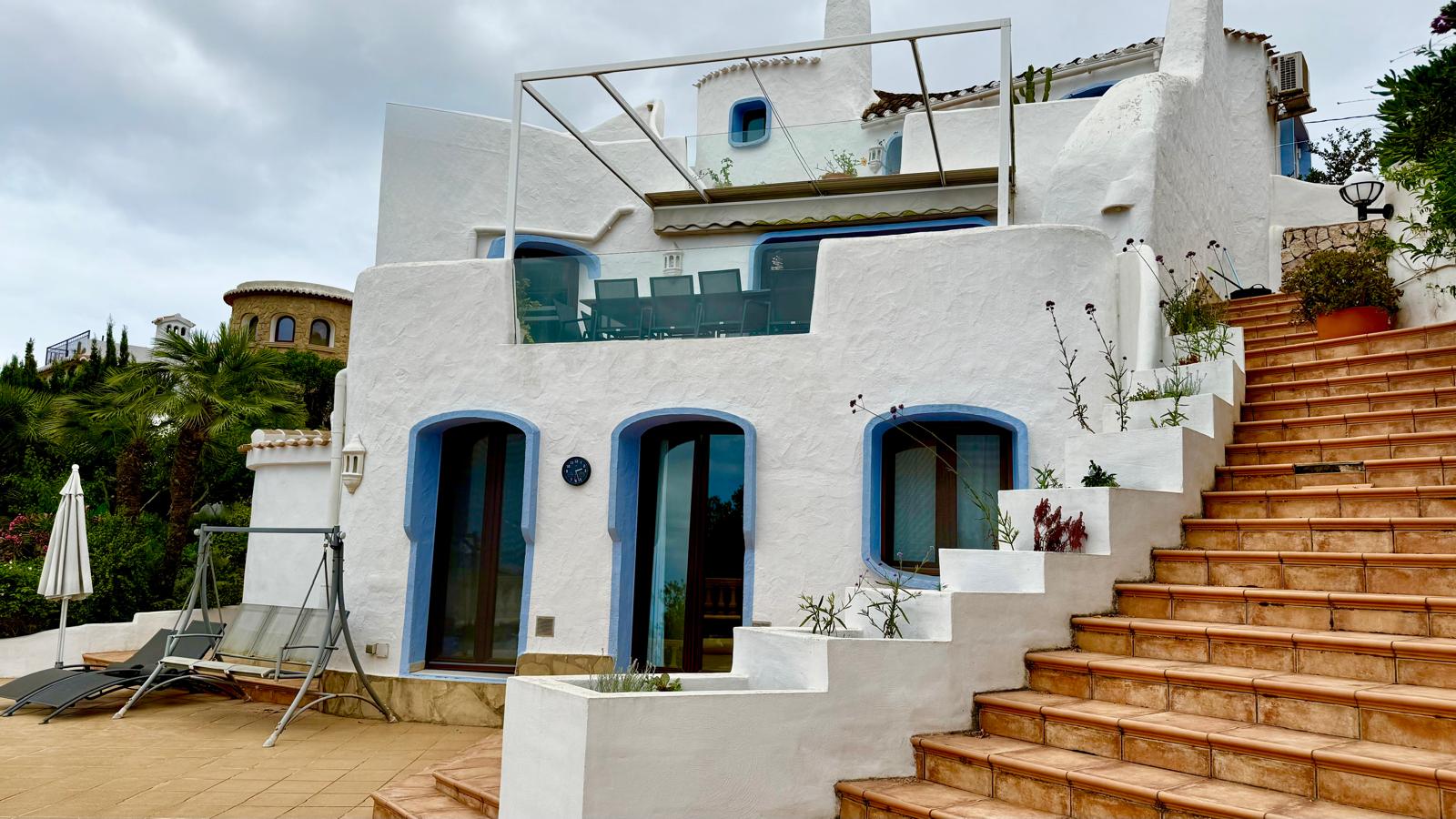 Außenansicht im ibizenkischen Stil mit weißen Wänden und blauen Akzenten. Terrakotta-Treppen verbinden die verschiedenen Ebenen und Terrassen mit Glasgeländern.