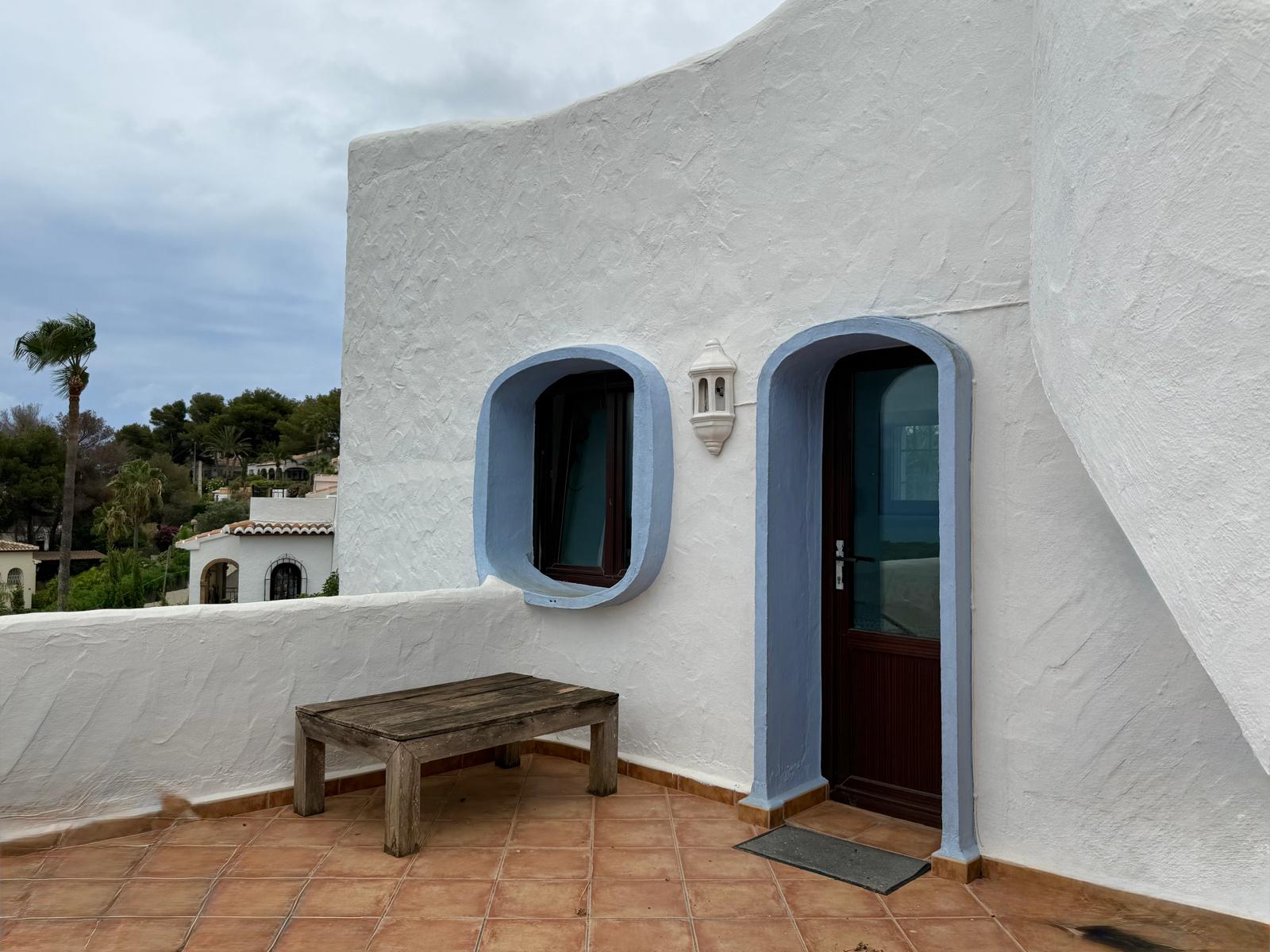 Außenfassade im ibizenkischen Stil mit weißem Strukturputz, dunklen Holzrahmen und blau abgesetzten Rundbögen. Die Terrasse ist mit Terrakottafliesen und einer rustikalen Holzbank ausgestattet.