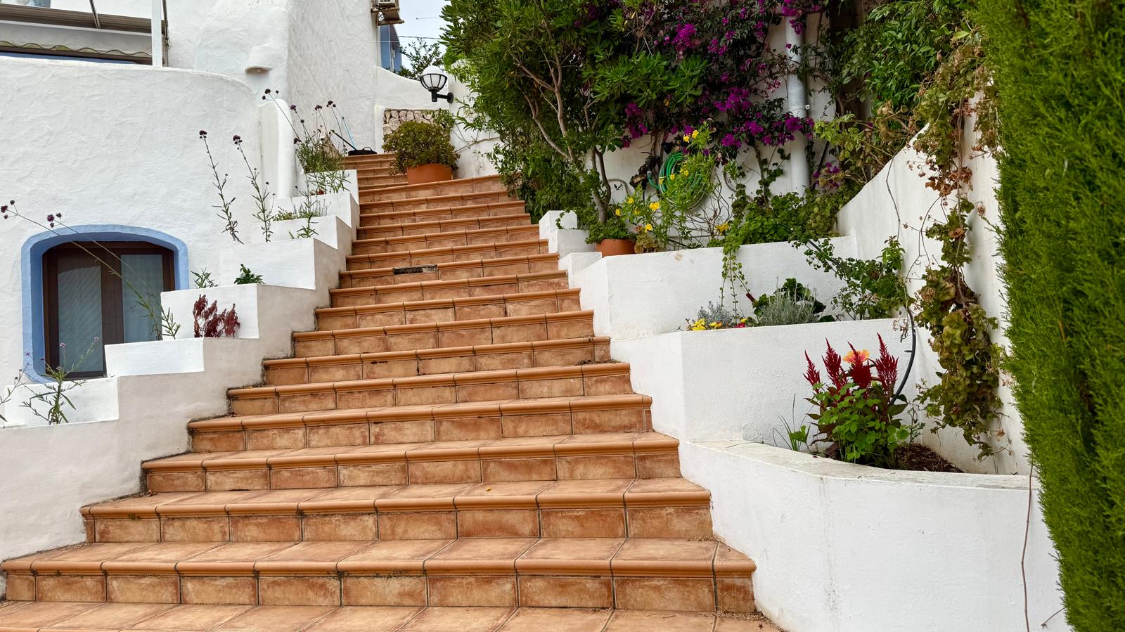 Außenliegende Treppe mit Terrakotta-Fliesen, die die Ebenen verbindet, gesäumt von weiß verputzten Pflanzkübeln mit mediterraner Bepflanzung und Bougainvillea.