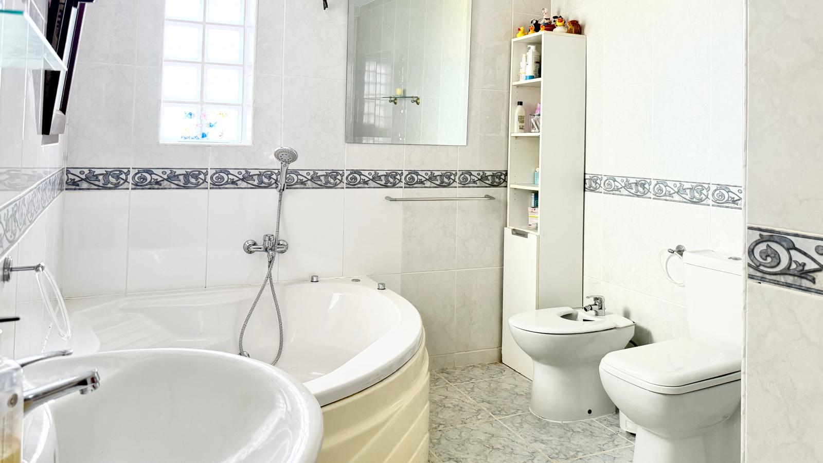 Badezimmer mit einer großen Eck-Hydromassage-Badewanne, Bidet und WC, ausgestattet mit weißen Wandfliesen und einer klassischen dekorativen Bordüre.