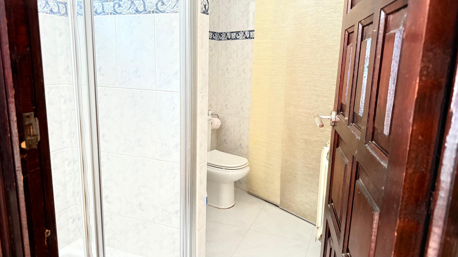 Badezimmer mit Glasduschkabine, WC und hell gefliesten Wänden mit blauer Zierborte. Im Vordergrund befindet sich eine dunkle Holztür.