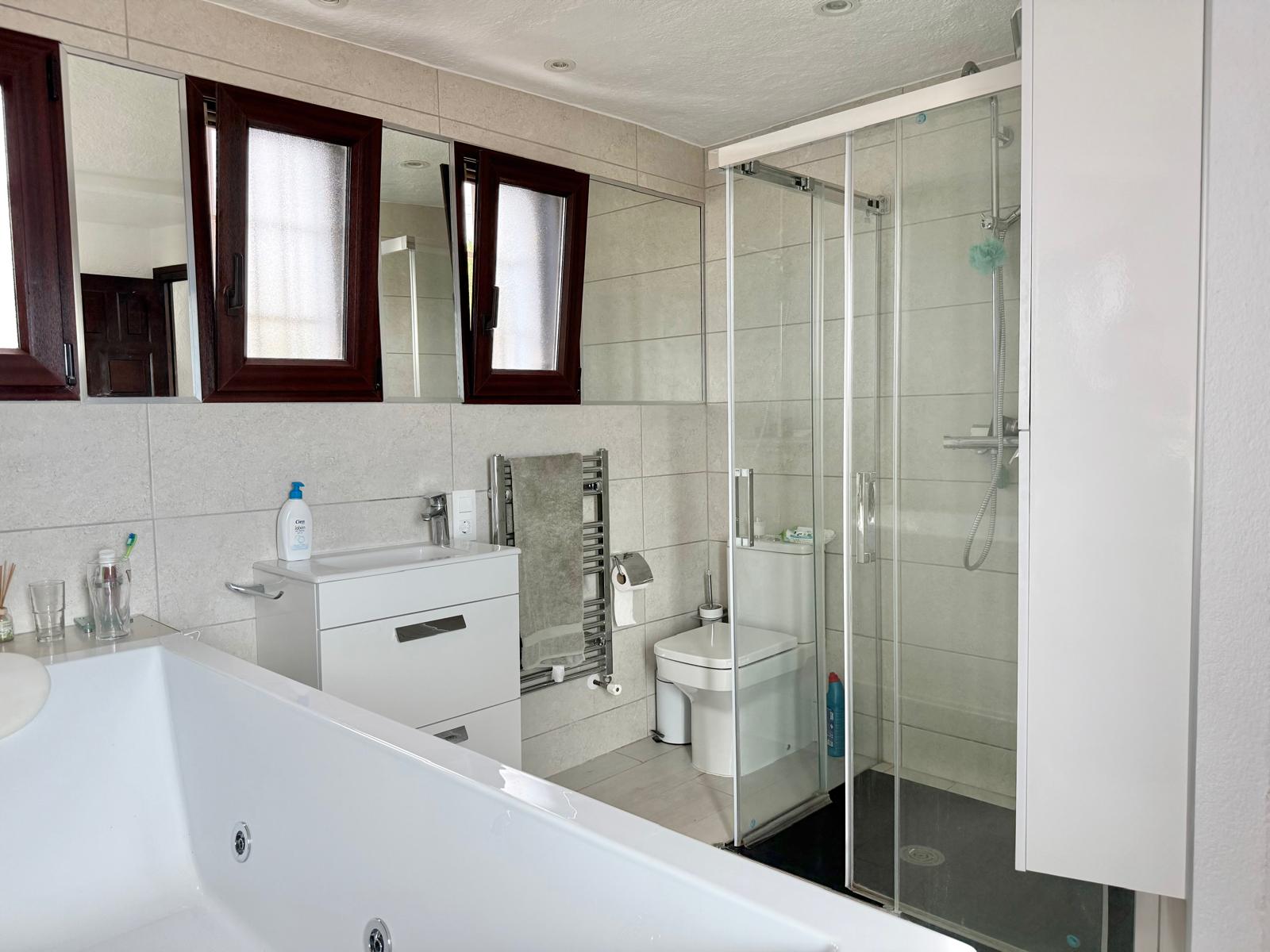 Badezimmer mit Hydromassage-Badewanne, einer begehbaren Glasdusche, weißem Waschtisch und Handtuchheizkörper vor hellen Fliesenwänden.