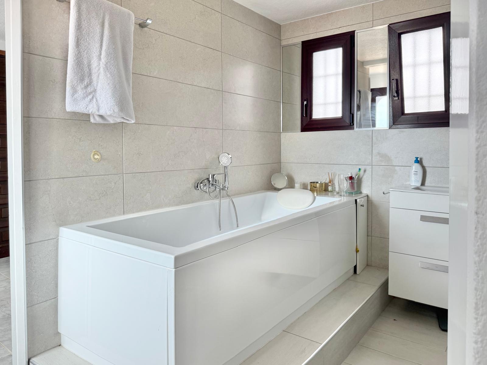 Badezimmer mit weißer Badewanne, neutralen Wandfliesen und einem weißen Waschtisch. Dunkle Fensterrahmen und verchromte Armaturen sorgen für ein modernes Erscheinungsbild.