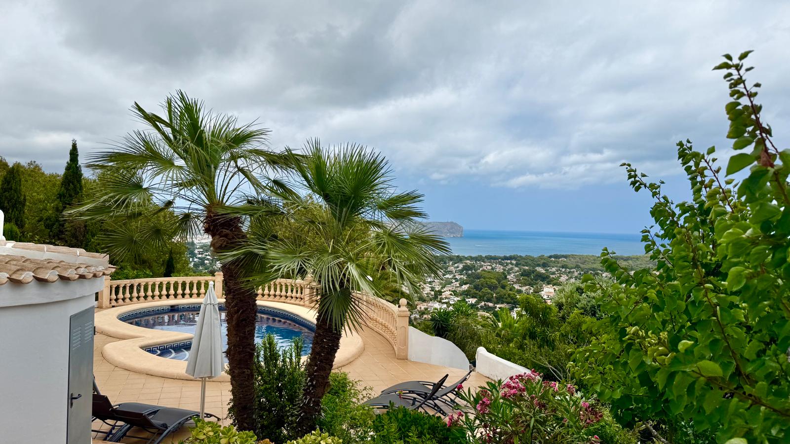 Erhöhter Blick auf den geschwungenen Pool und den mediterranen Garten mit Panoramasicht auf die Bucht von Jávea und das Meer hinter Palmen.