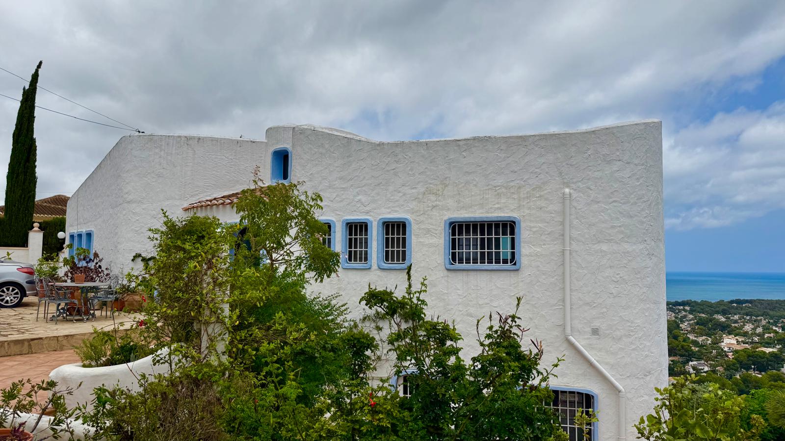 Fassade im Ibiza-Stil mit weiß verputzten Wänden, blauen Fensterrahmen und Gittern. Die Steinterrasse bietet einen weiten Blick auf das Meer und die umliegende Natur.