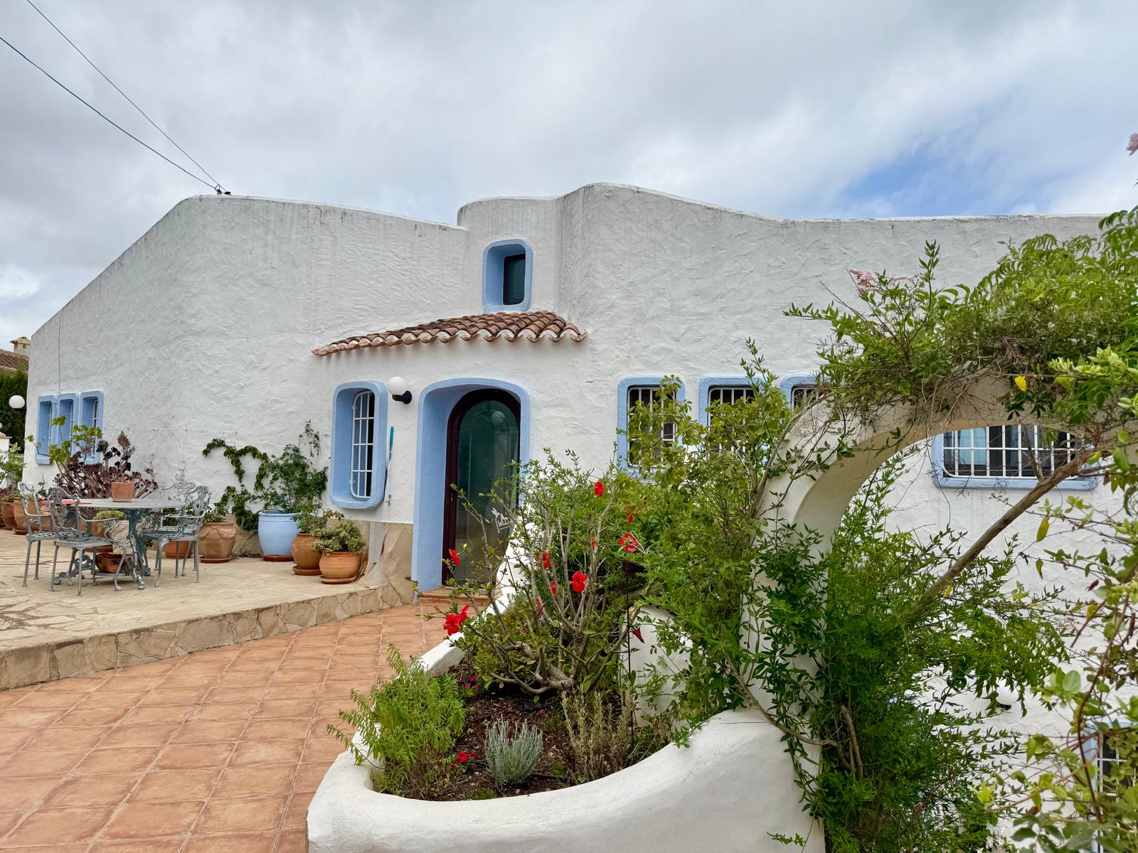 Fassade im ibizenkischen Stil mit organisch geformten weißen Wänden, blauen Fensterrahmen und einer Terrakotta-Terrasse mit mediterraner Bepflanzung.