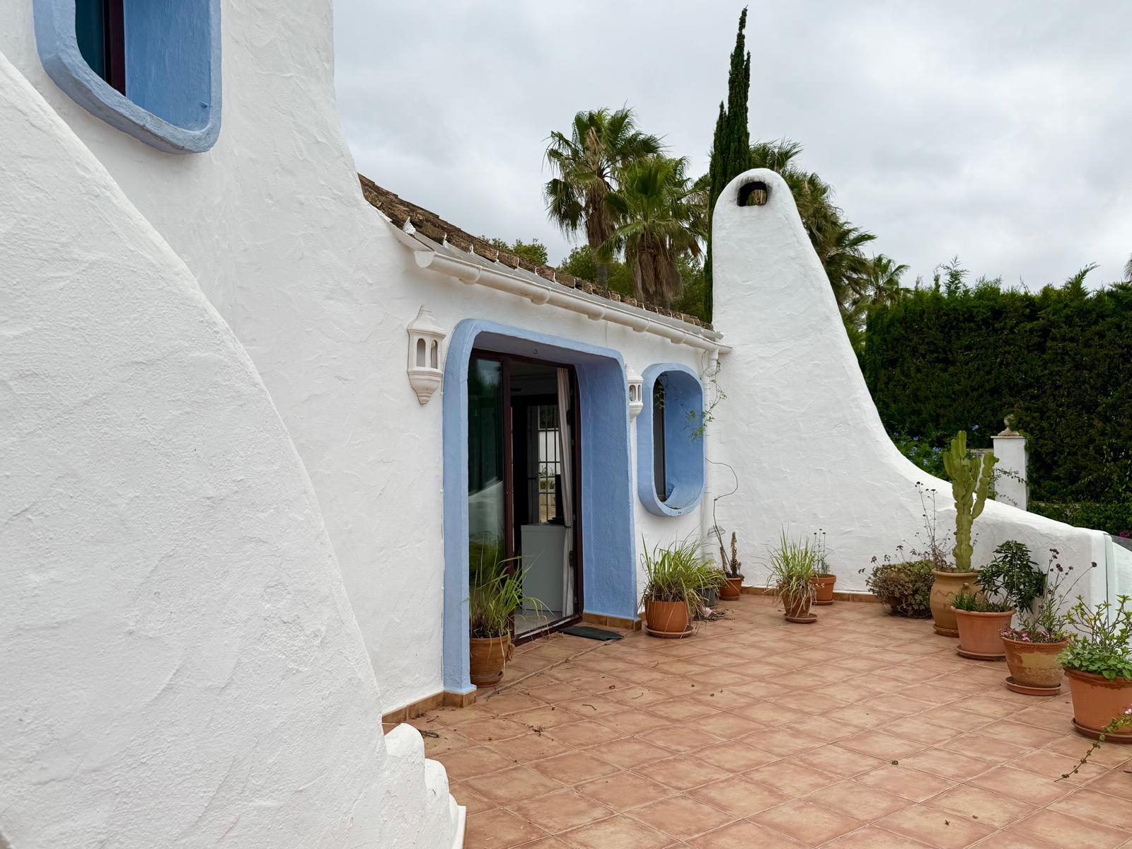 Fassade im ibizenkischen Stil mit organischen weißen Wänden, blauen Fensterrahmen und markantem Kamin. Die Terrasse mit Terrakotta-Fliesen ist mit mediterranen Kübelpflanzen bestückt.