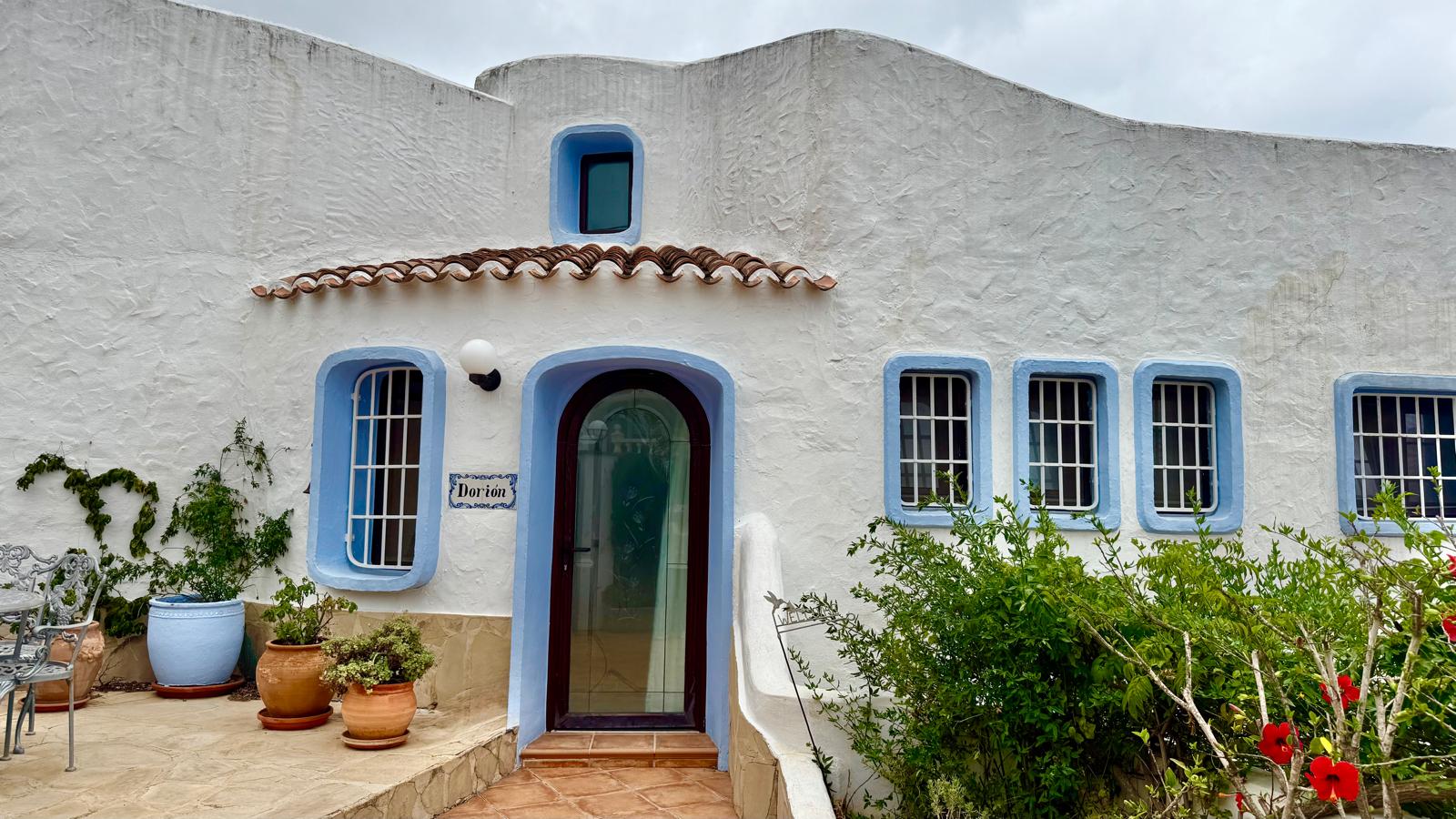 Fassade im ibizenkischen Stil mit weißen Wänden, blauen Fensterumrandungen und einer bogenförmigen Glastür. Terrasse mit Terrakottafliesen und mediterranen Kübelpflanzen.