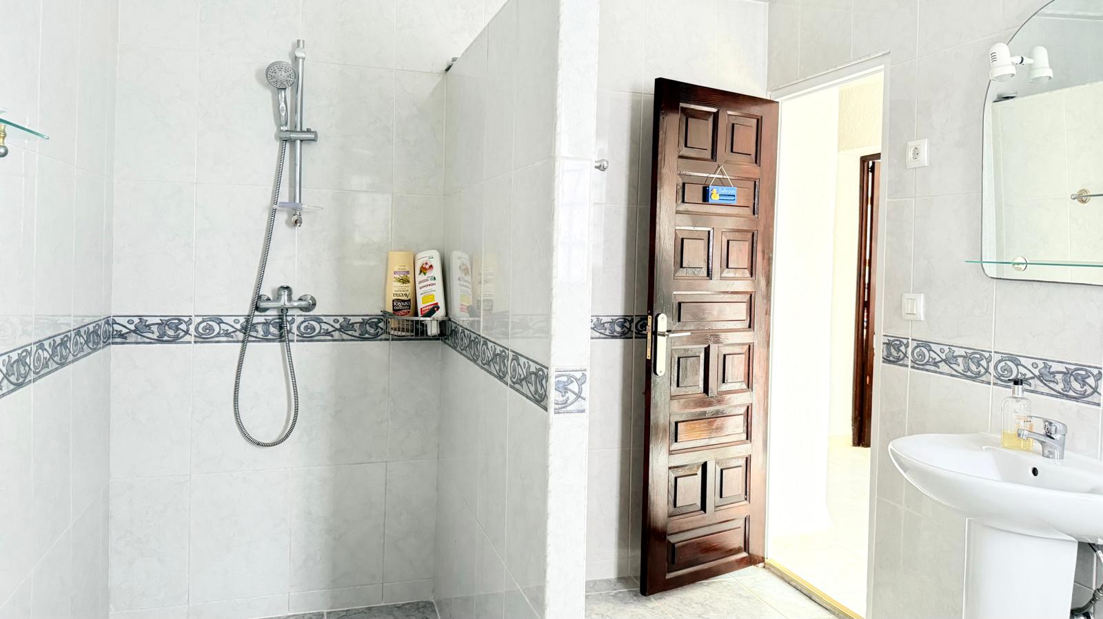 Funktionales Badezimmer mit begehbarer Dusche, dekorativer Fliesenfuge, einer geschnitzten Massivholztür und einem Säulenwaschbecken mit beleuchtetem Spiegel.