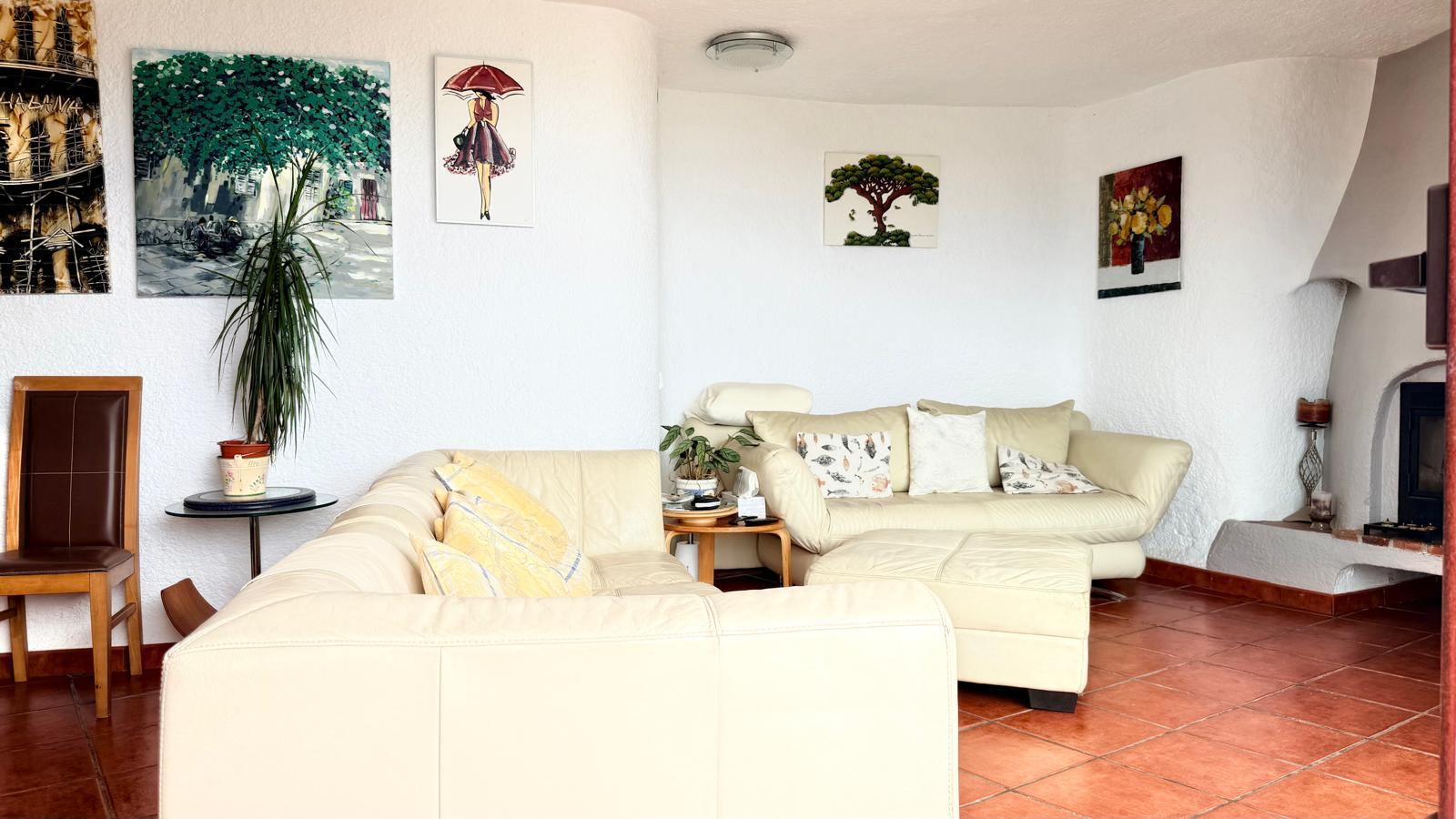 Gemütlicher Wohnbereich mit Holzkamin, weißen strukturierten Wänden im Ibiza-Stil, Terrakottaboden und hellen Ledersofas in einer geschwungenen Architektur.