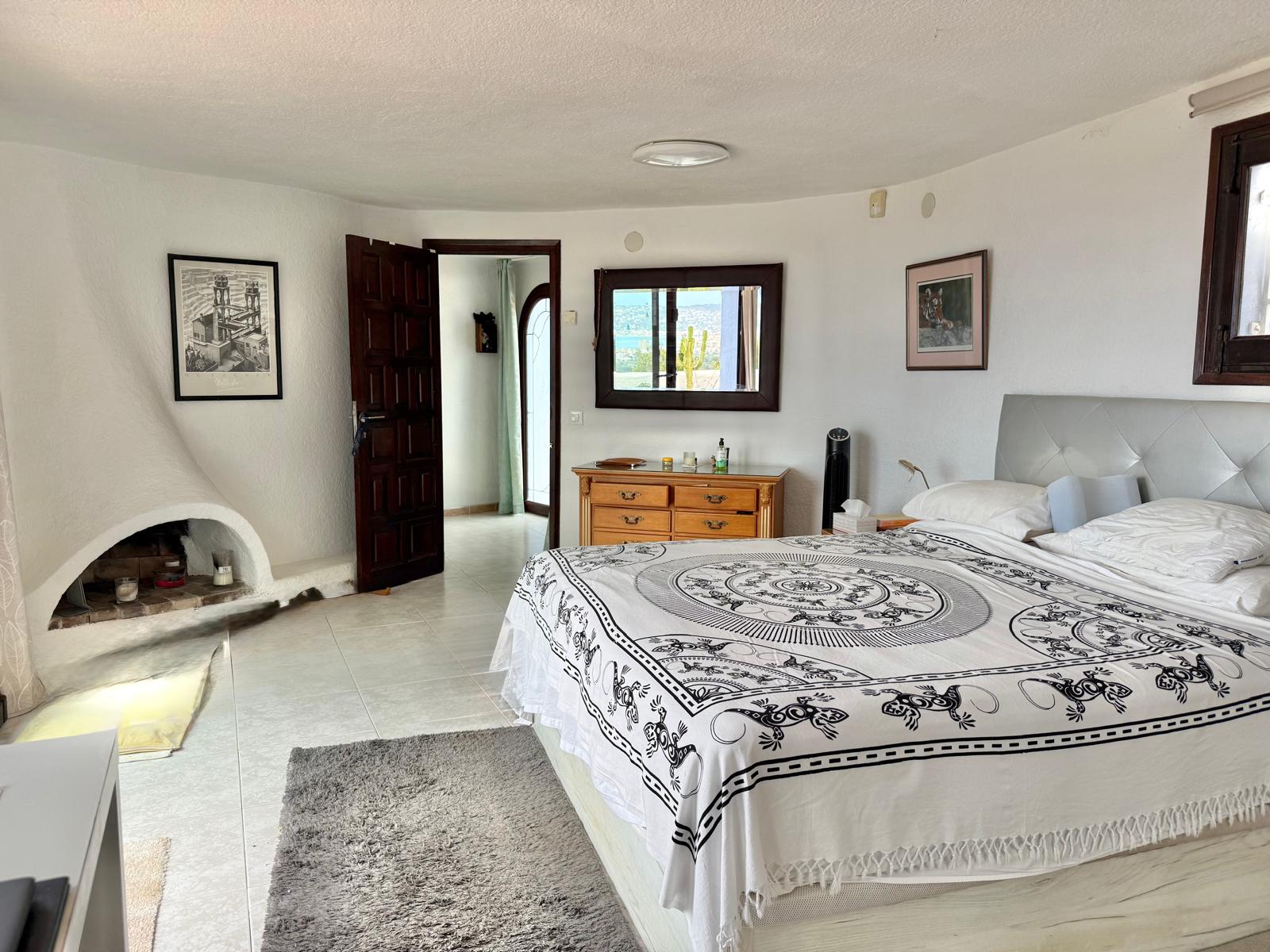 Hauptschlafzimmer im Ibiza-Stil mit geschwungenem gemauertem Kamin, Fliesenboden und Spiegel, der den Panoramablick auf das Meer einfängt.