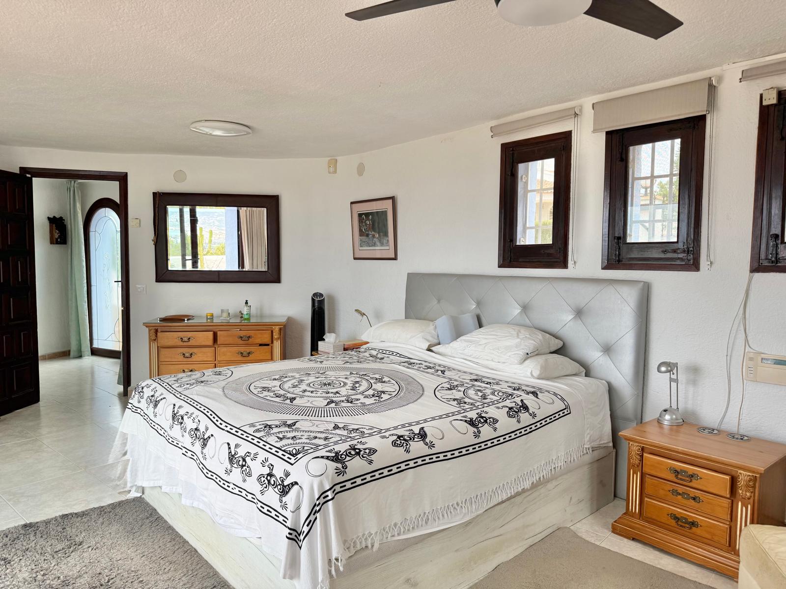 Hauptschlafzimmer mit weißen Wänden, dunklen Holzfensterrahmen und Deckenventilator. Es bietet klassische Holzmöbel und einen Durchgang zum angrenzenden Ankleidebereich.