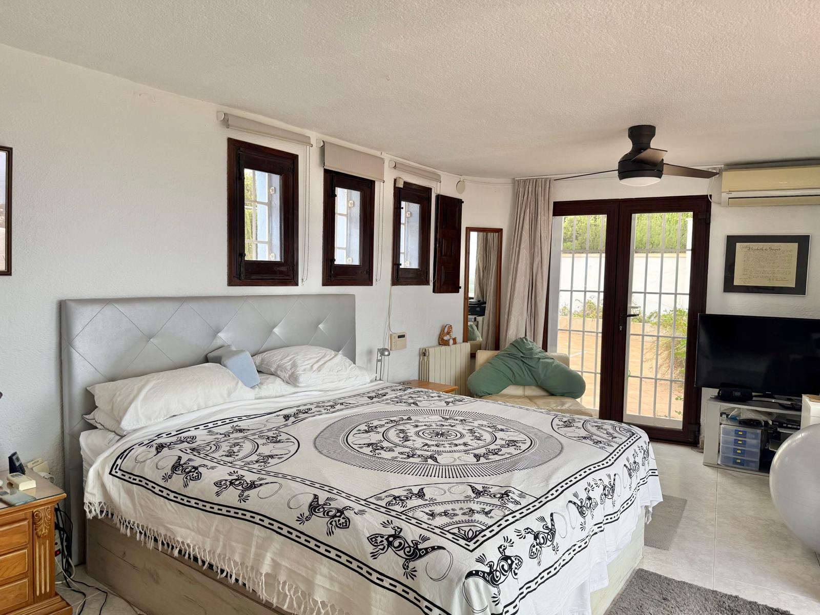 Hell gestaltetes Hauptschlafzimmer mit dunklen Holzfensterrahmen, Deckenventilator und direktem Zugang zur Terrasse durch große Glastüren.