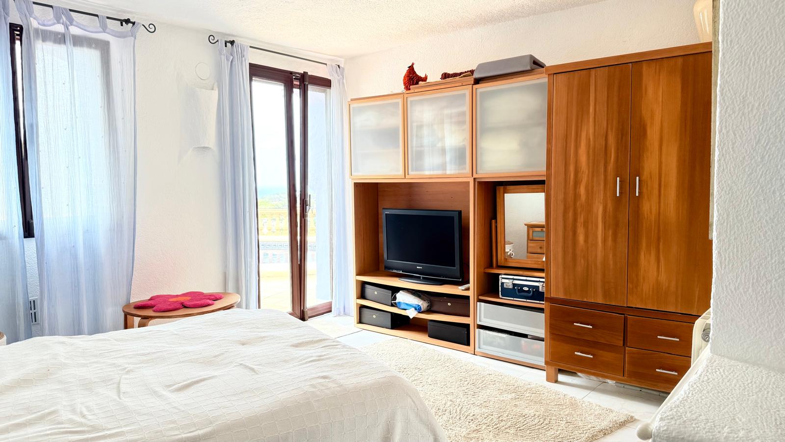 Helles Schlafzimmer mit integrierter Holzschrankwand, TV-Bereich und direktem Zugang zur Terrasse mit weitem Blick auf das Meer.
