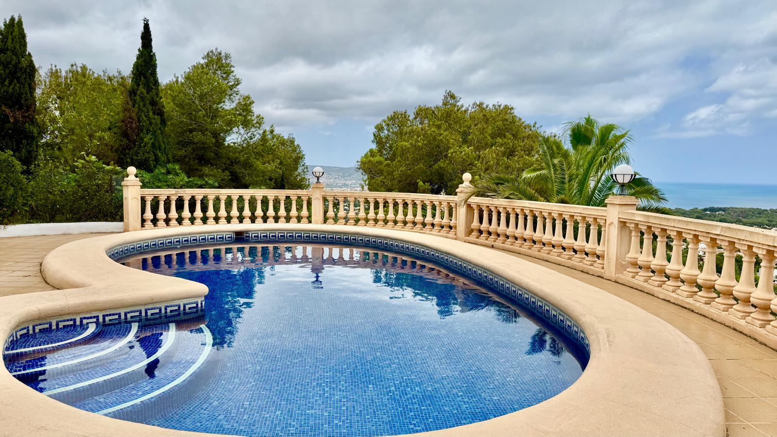 Privater, nierenförmiger Pool mit blauen Mosaikfliesen, umgeben von einer Steinterrasse und klassischer Balustrade mit Panoramablick auf das Meer und Pinien.