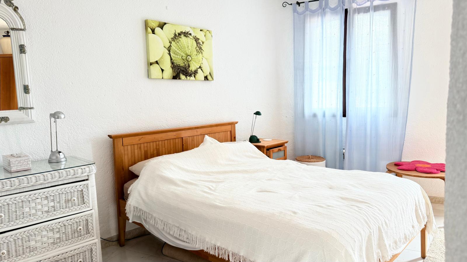 Schlafzimmer mit Holzbettrahmen, weißer Rattankommode und strukturierten Wänden. Natürliches Licht fällt durch ein Fenster mit hellblauen Vorhängen in den Raum.