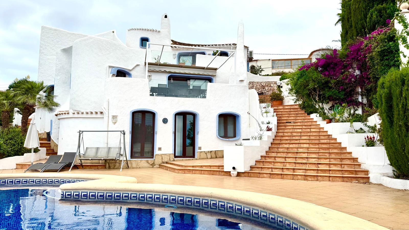 Villa im Ibiza-Stil mit weißen Rundbögen, blauen Akzenten, einem großen Pool und einer Außentreppe aus Terrakotta-Fliesen inmitten mediterraner Bepflanzung.