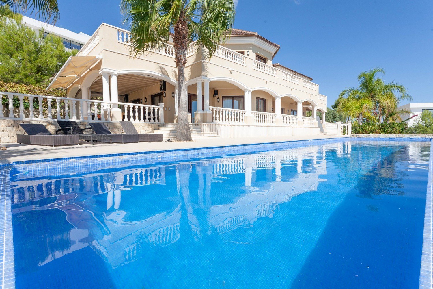 Blick auf den blau gefliesten "Infinity"-Pool und die Fassade der mediterranen Villa mit Säulen.