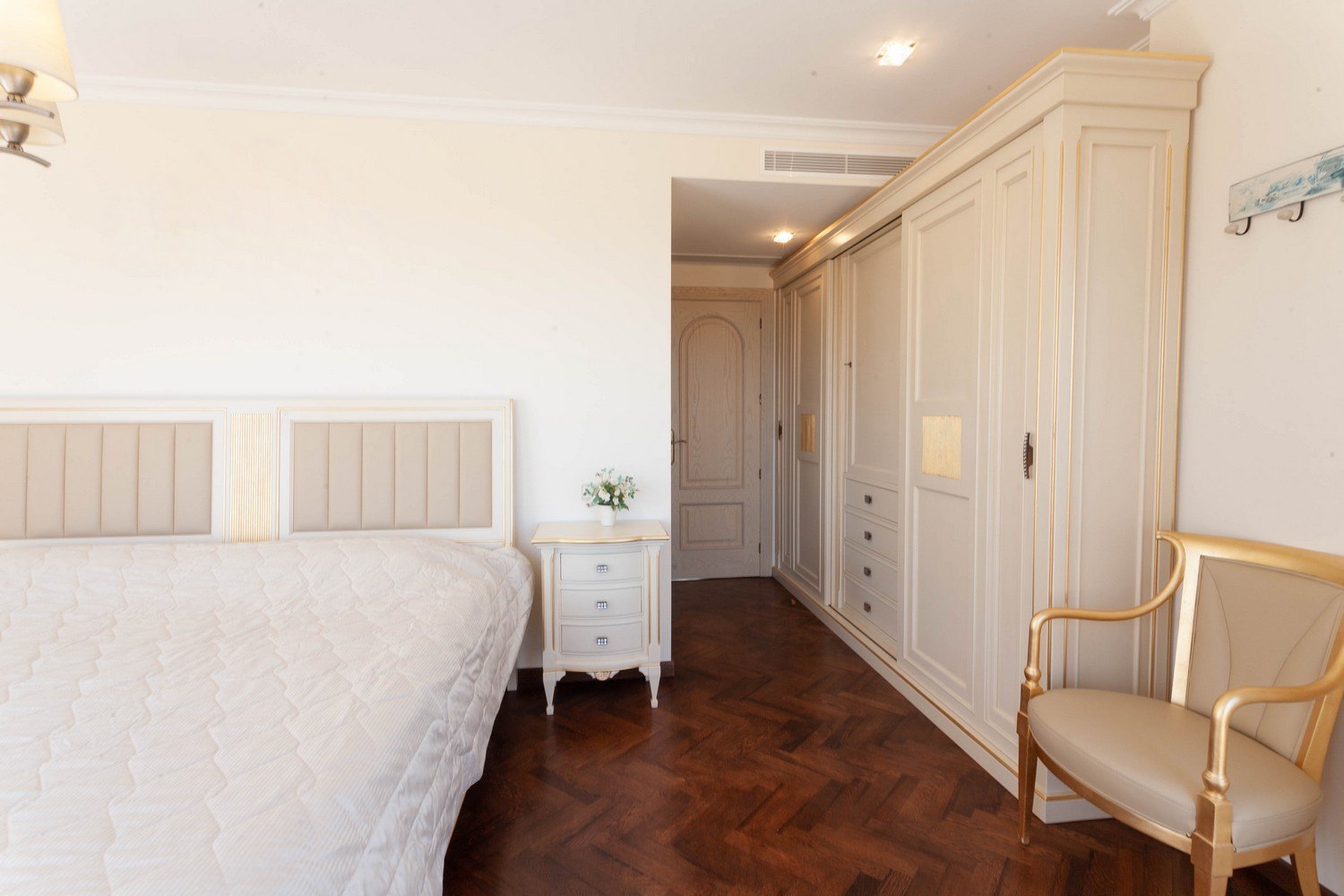 Schlafzimmer mit Bett mit gepolstertem Kopfteil, Einbauschrank aus hellem Holz und dunklem Parkettboden.
