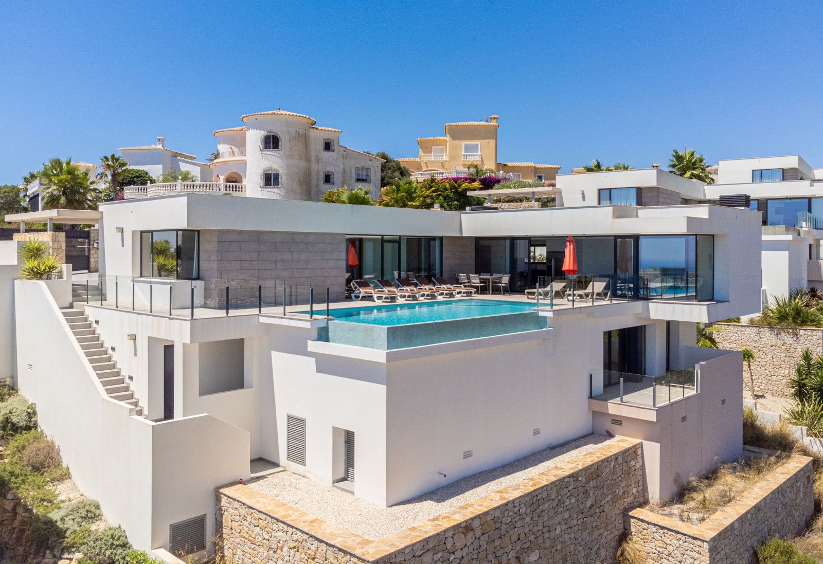 Außenansicht der modernen Villa mit erhöhtem Infinity-Pool, weißer Fassade mit Steinelementen und großen Glasfronten zu den Terrassen.