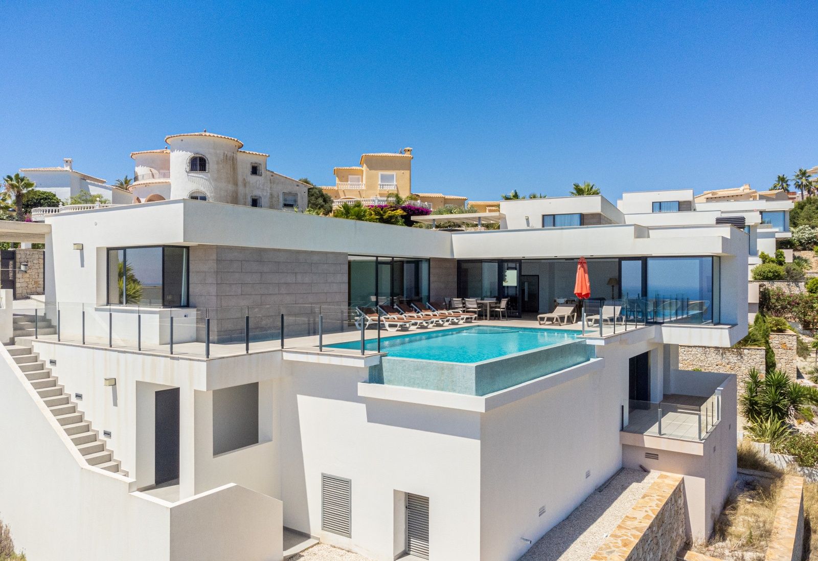 Außenansicht der modernen Villa mit Infinity-Pool, großzügiger Sonnenterrasse und weißer Fassade mit grauen Steinakzenten.