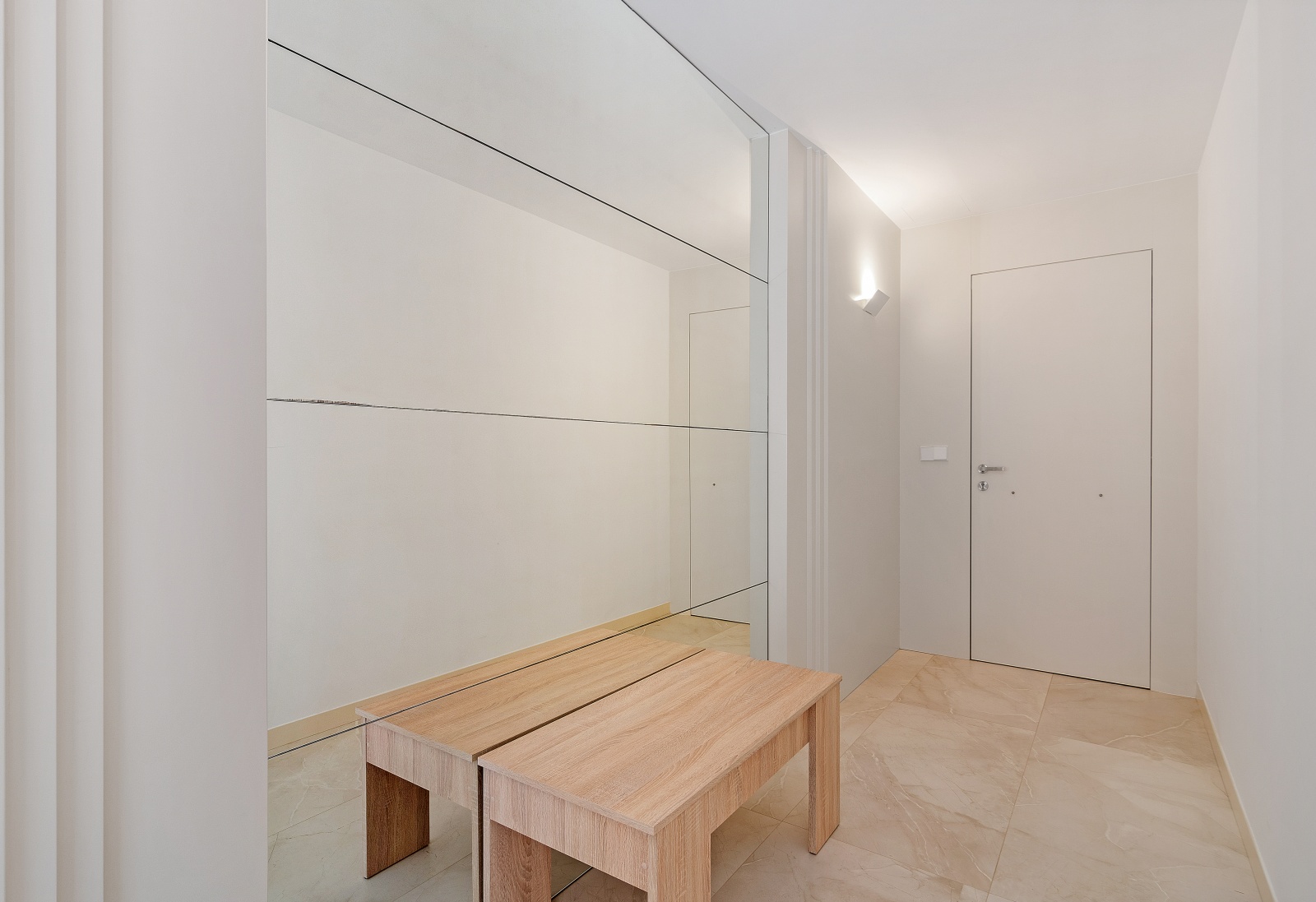 Eingangsbereich mit großem Wandspiegel, einer hellen Holzbank und cremefarbenen Feinsteinzeugfliesen. Minimalistisches Design mit schlichter weißer Tür und Wandbeleuchtung.