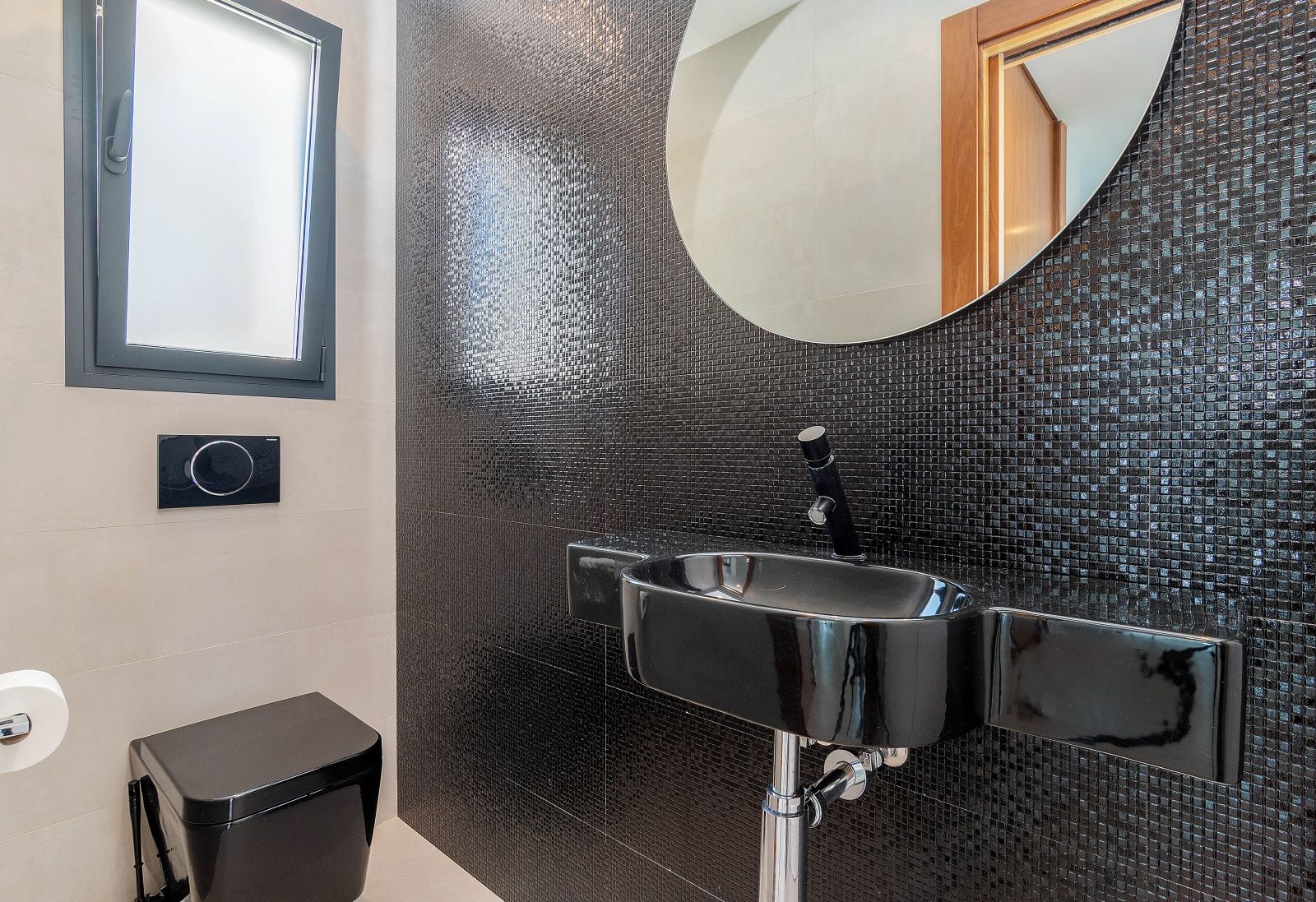 Gäste-WC mit einer Akzentwand aus schwarzem Mosaik, passendem schwarzen Waschbecken und Wand-WC. Ein runder Spiegel und ein mattiertes Fenster sorgen für Licht.