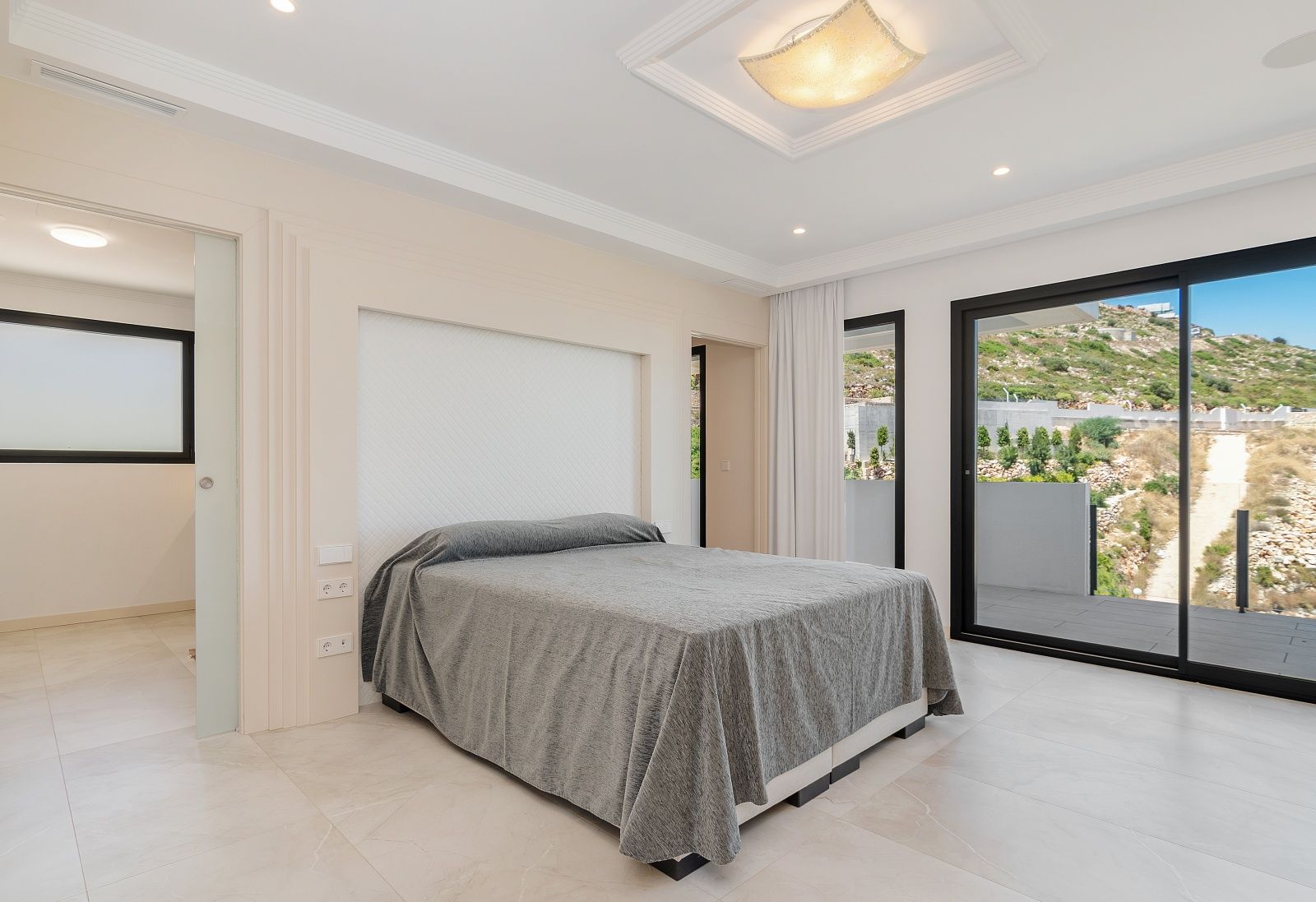 Helles Schlafzimmer mit großen Glasschiebetüren zur Terrasse, integrierter Kopfteil-Nische und eleganter Deckenbeleuchtung mit Zierleisten.