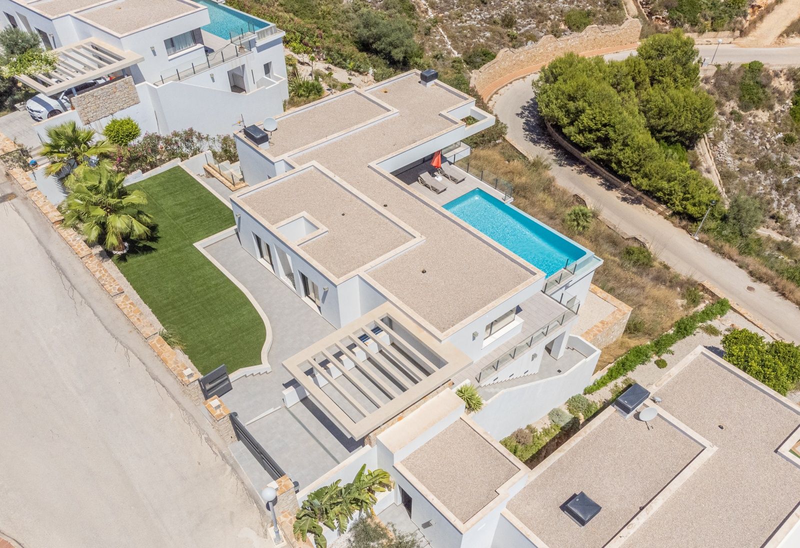Luftaufnahme der modernen Villa mit geradliniger Architektur, Infinity-Pool, flachen Kiesdächern und einer gepflegten Kunstrasenfläche.