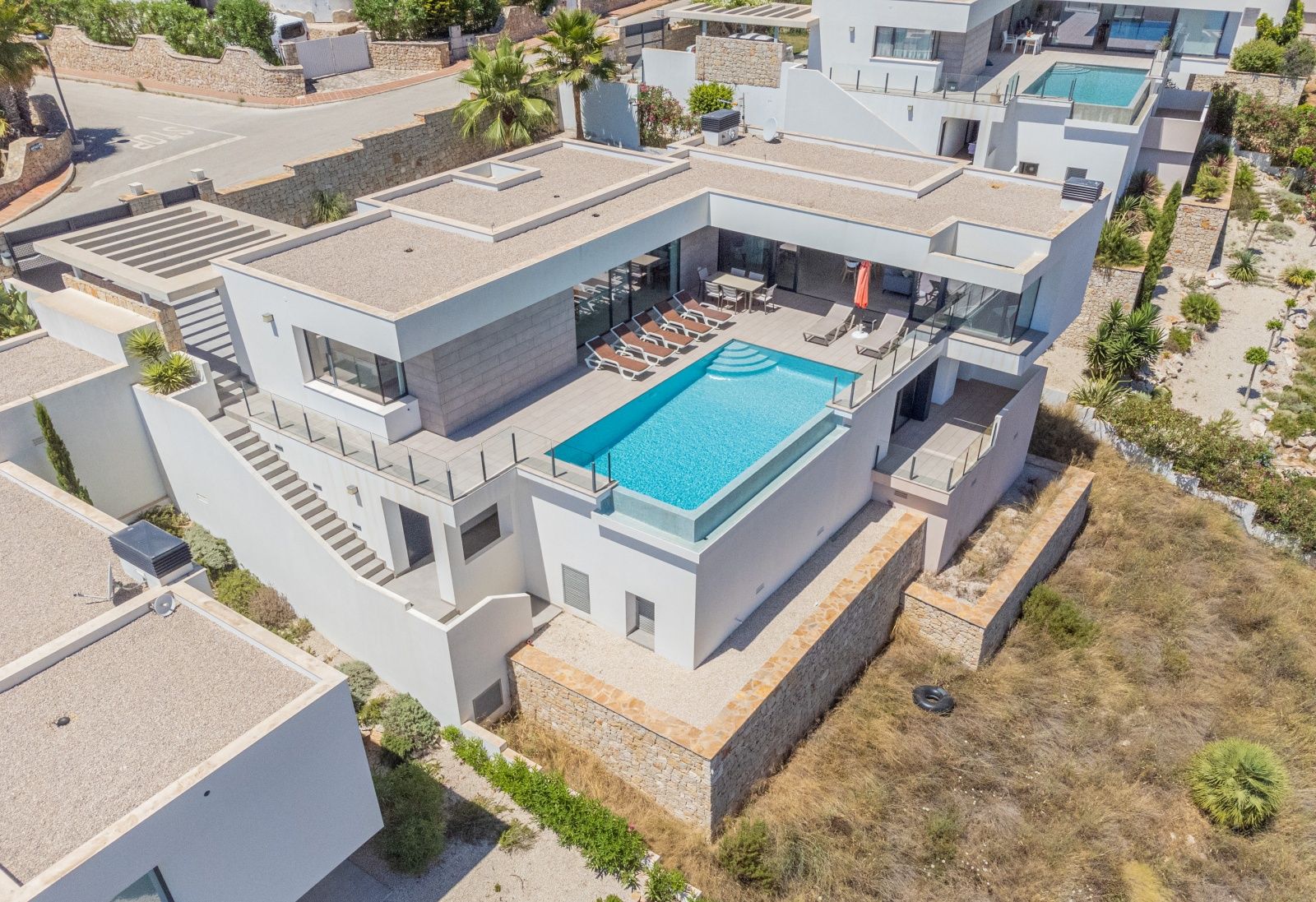 Luftaufnahme der modernen Villa mit Infinity-Pool und Glaswand, weitläufiger Sonnenterrasse mit Liegen und einem Essbereich im Freien in minimalistischem Design.