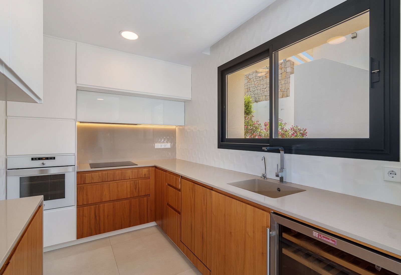 Moderne Küche mit Holz- und weißen Schränken, Steinarbeitsplatte, integrierten Geräten, Weinkühlschrank und großem Fenster mit schwarzem Rahmen.