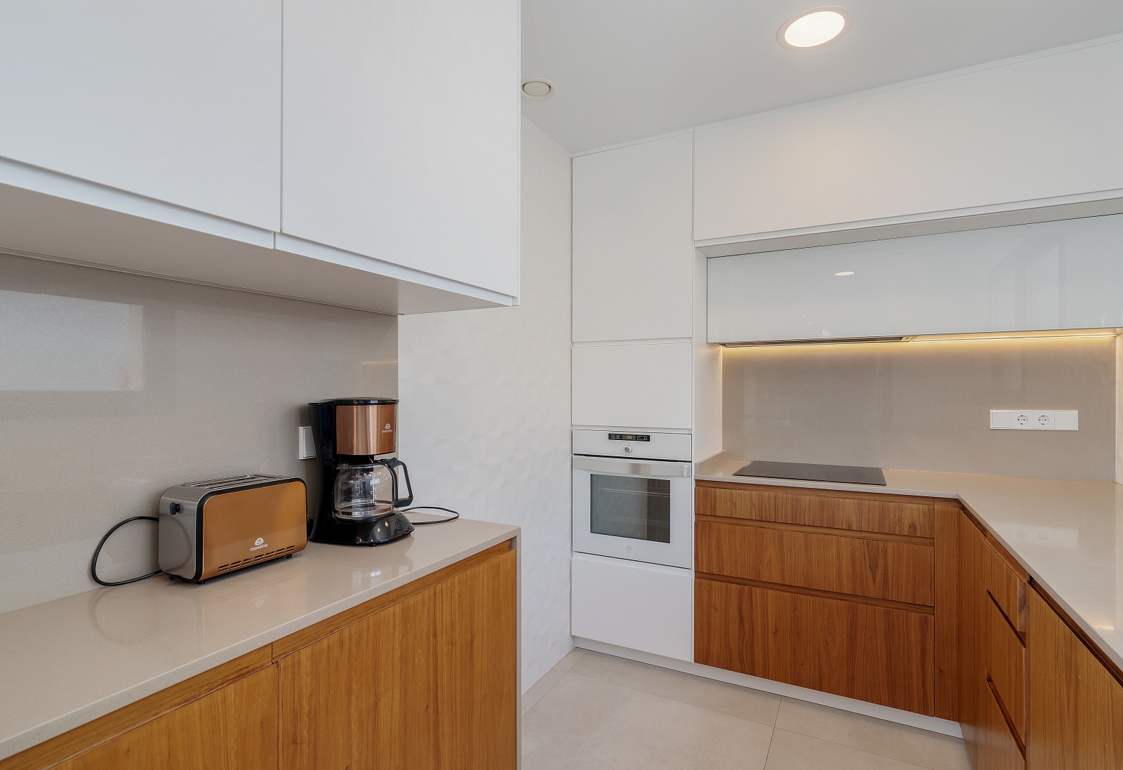 Moderne Küche mit weißen Oberschränken, Holzunterschränken, integriertem Backofen, Induktionskochfeld und Arbeitsplatte aus Quarzstein mit LED-Beleuchtung.
