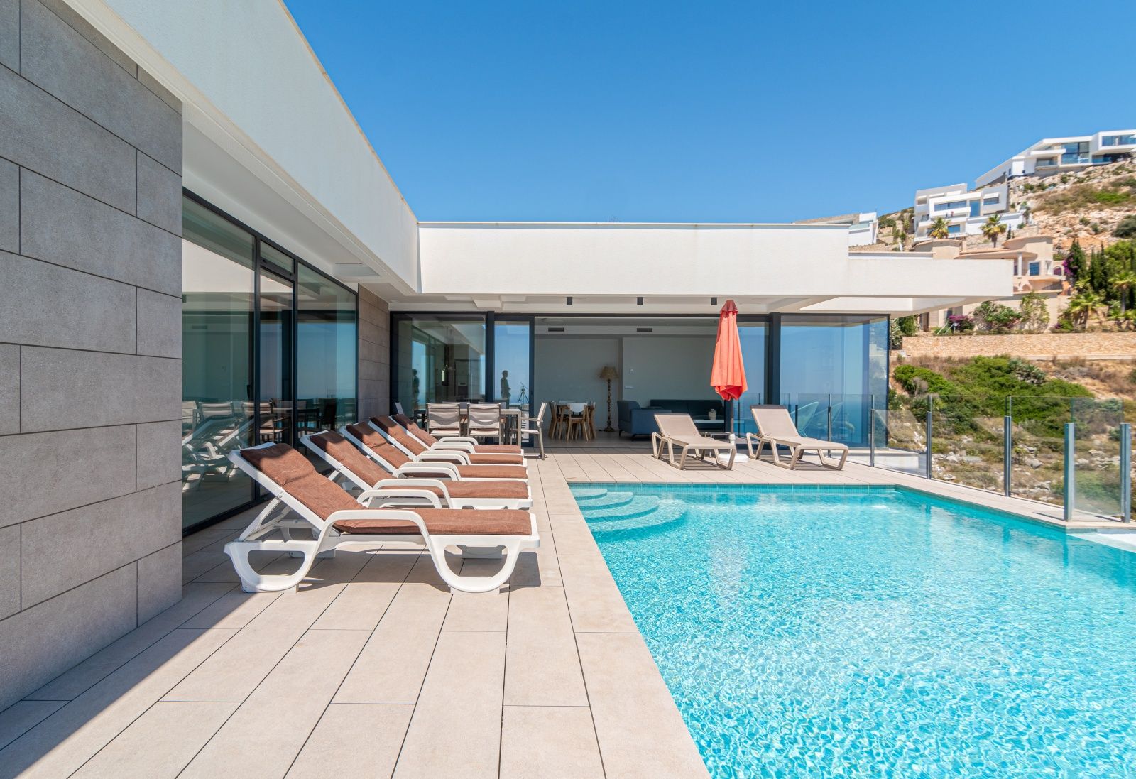 Moderne Terrasse mit Infinity-Pool, Sonnenliegen und großen Glasfronten, die den Innenbereich nahtlos mit dem Außenbereich und dem Solarium verbinden.