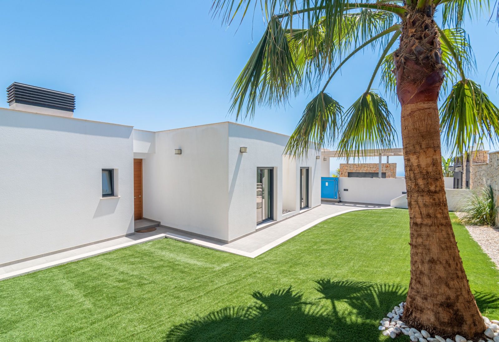 Moderne weiße Villa mit gepflegtem Kunstrasen, einer Palme im Vordergrund und einer Eingangstür aus Holz unter strahlend blauem Himmel.