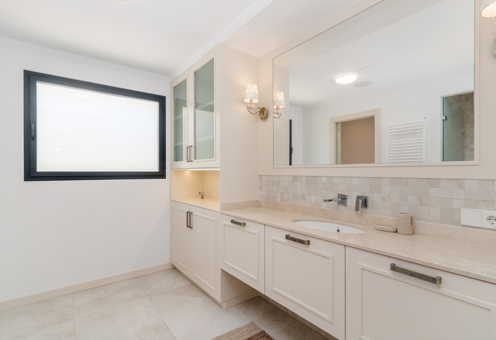 Modernes Badezimmer mit cremefarbenem Waschtisch, Natursteinplatte, Einbauschränken mit Glasfronten und großem Spiegel mit eleganten Wandleuchten.