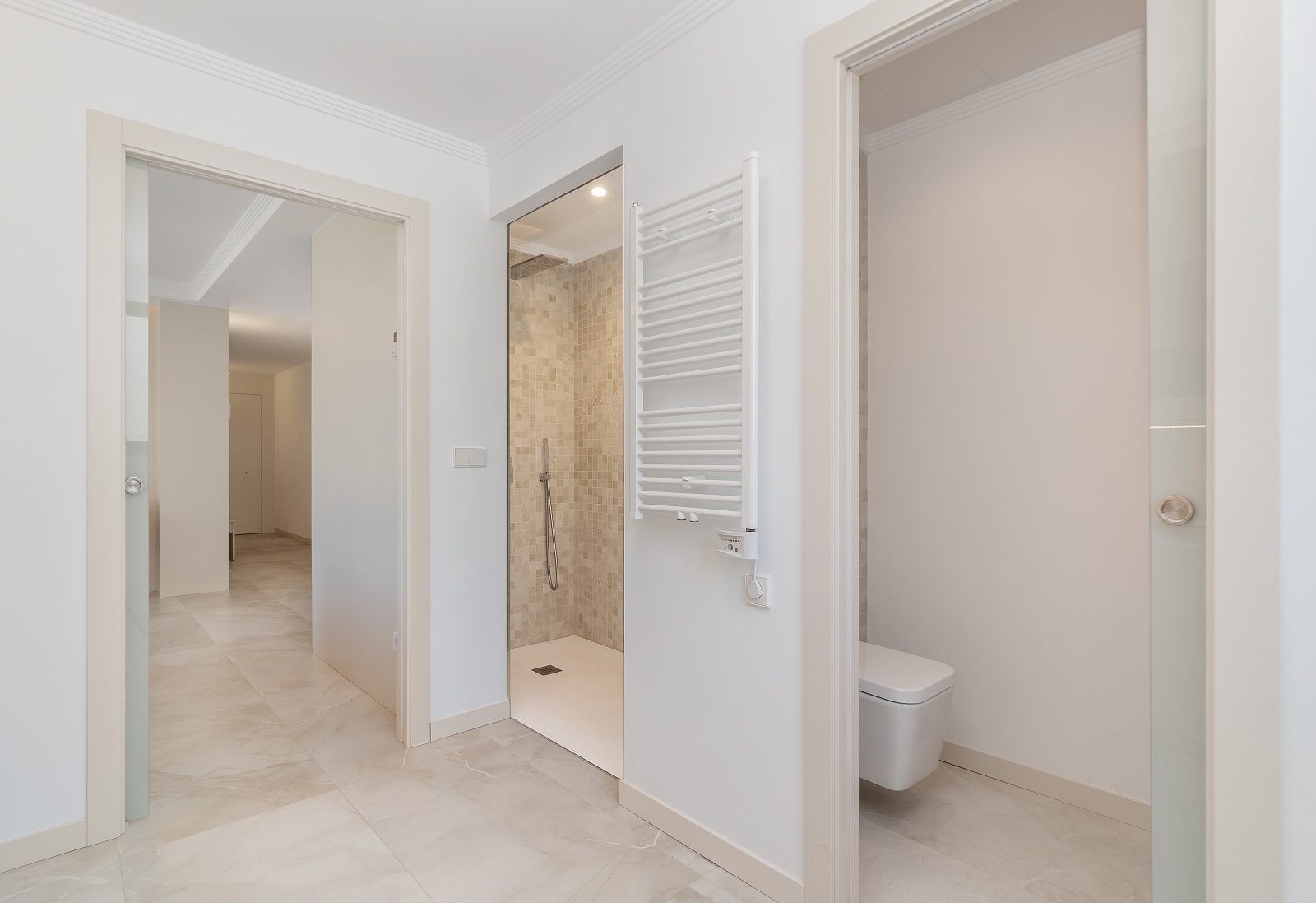 Modernes Badezimmer mit ebenerdiger Dusche in beigem Mosaik, weißem Handtuchheizkörper und wandhängendem WC. Hochwertige Fliesenböden und Glasschiebetüren.