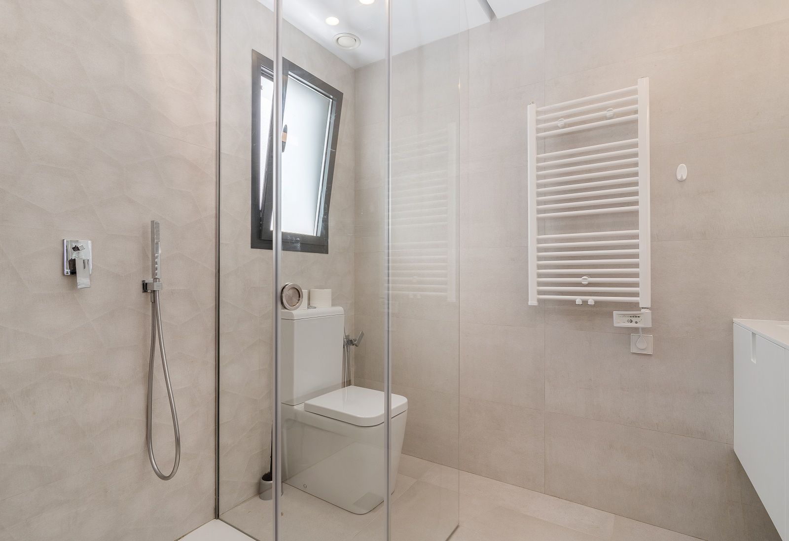 Modernes Badezimmer mit Glasduschkabine, strukturierten beigen Wandfliesen, weißem Handtuchheizkörper und integrierten Deckenspots.