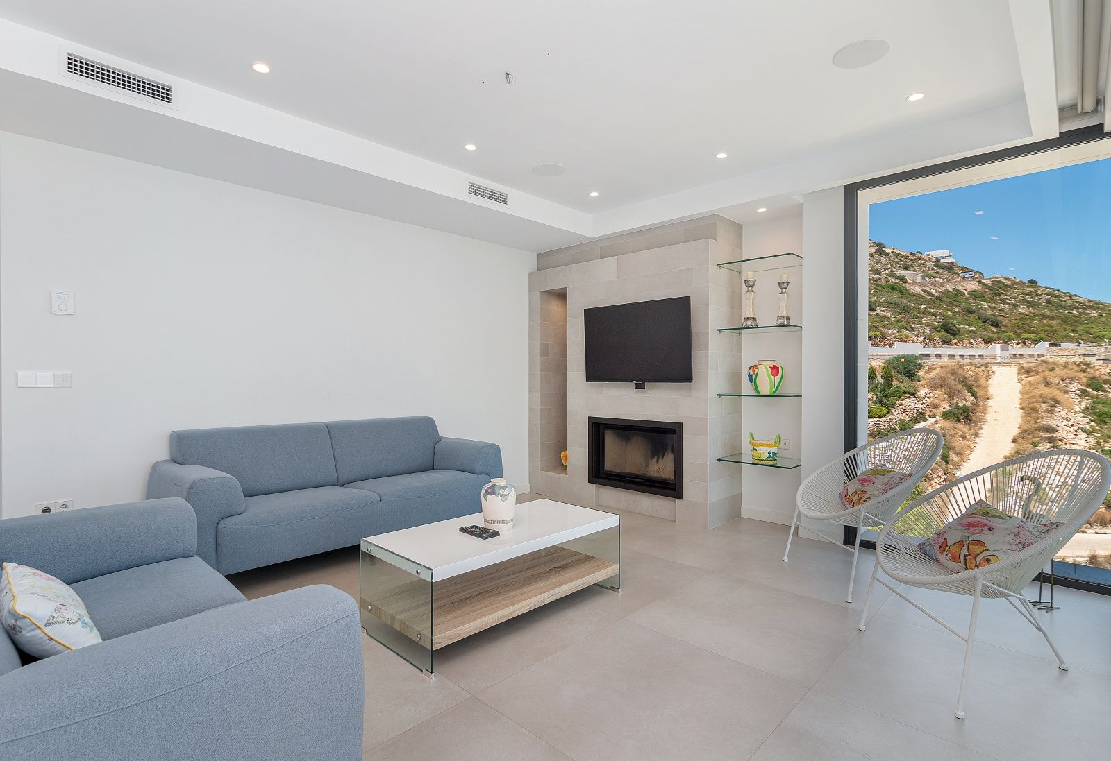 Modernes Wohnzimmer mit blauen Sofas, integriertem Kamin mit Steinverkleidung und bodentiefen Fenstern mit Blick auf die umliegende Hügellandschaft.