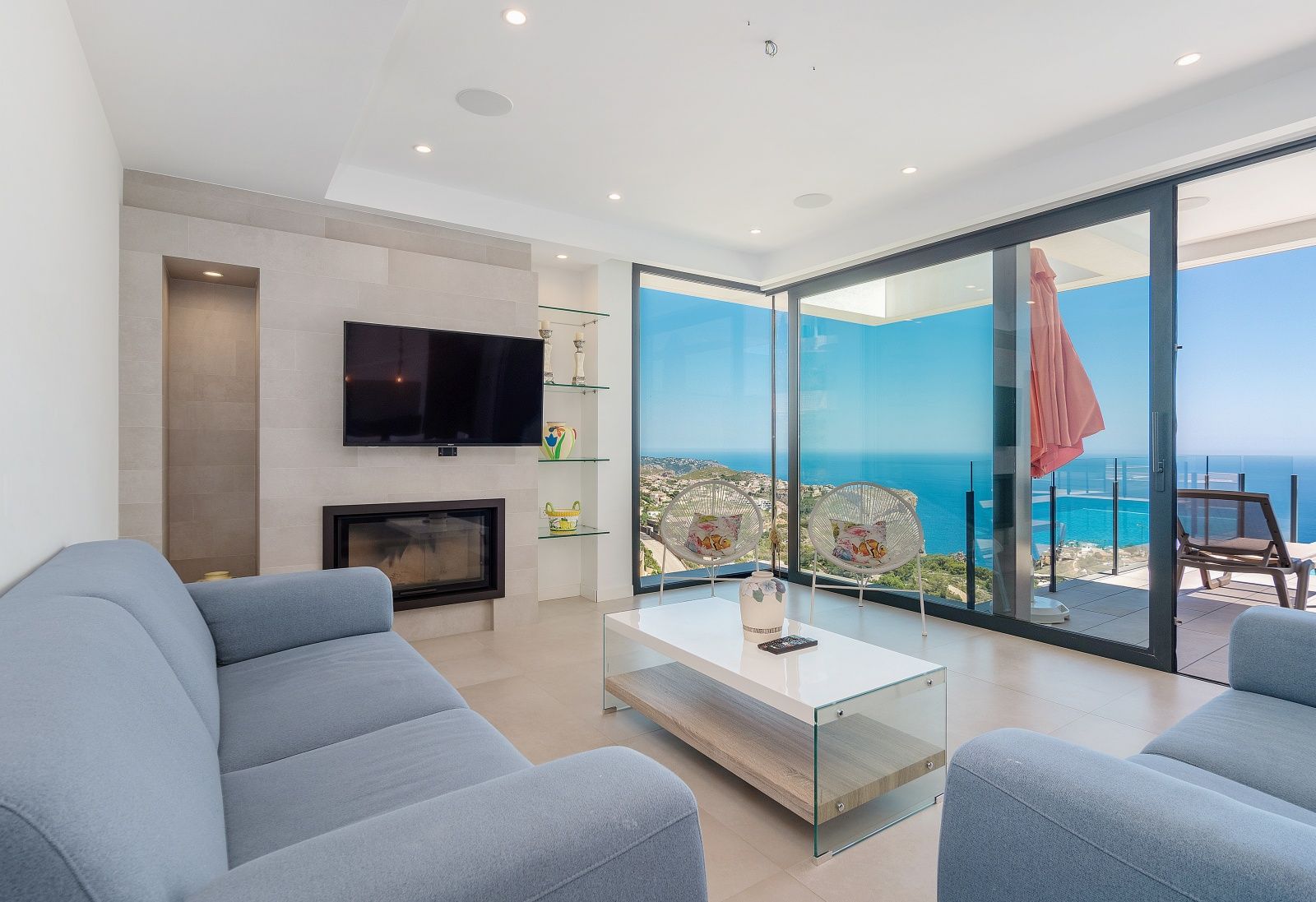 Modernes Wohnzimmer mit blauen Sofas, integriertem Kamin und bodentiefen Glasfronten mit Panoramablick auf das Mittelmeer.