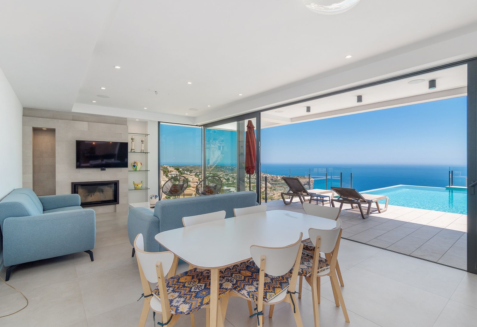 Offener Wohn- und Essbereich mit modernem Kamin und bodentiefen Glasfronten, die den Blick auf den Infinity-Pool und das Mittelmeer freigeben.