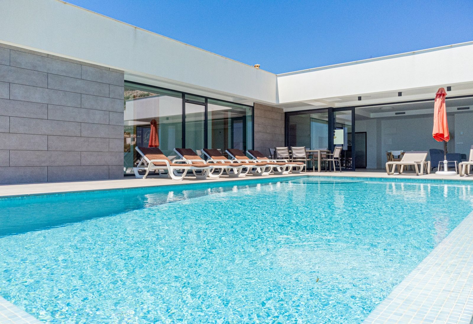 Rechteckiger Swimmingpool mit blauen Mosaikfliesen und großzügiger Sonnenterrasse. Die moderne Fassade kombiniert graue Steinpaneele mit großen Glasfronten.