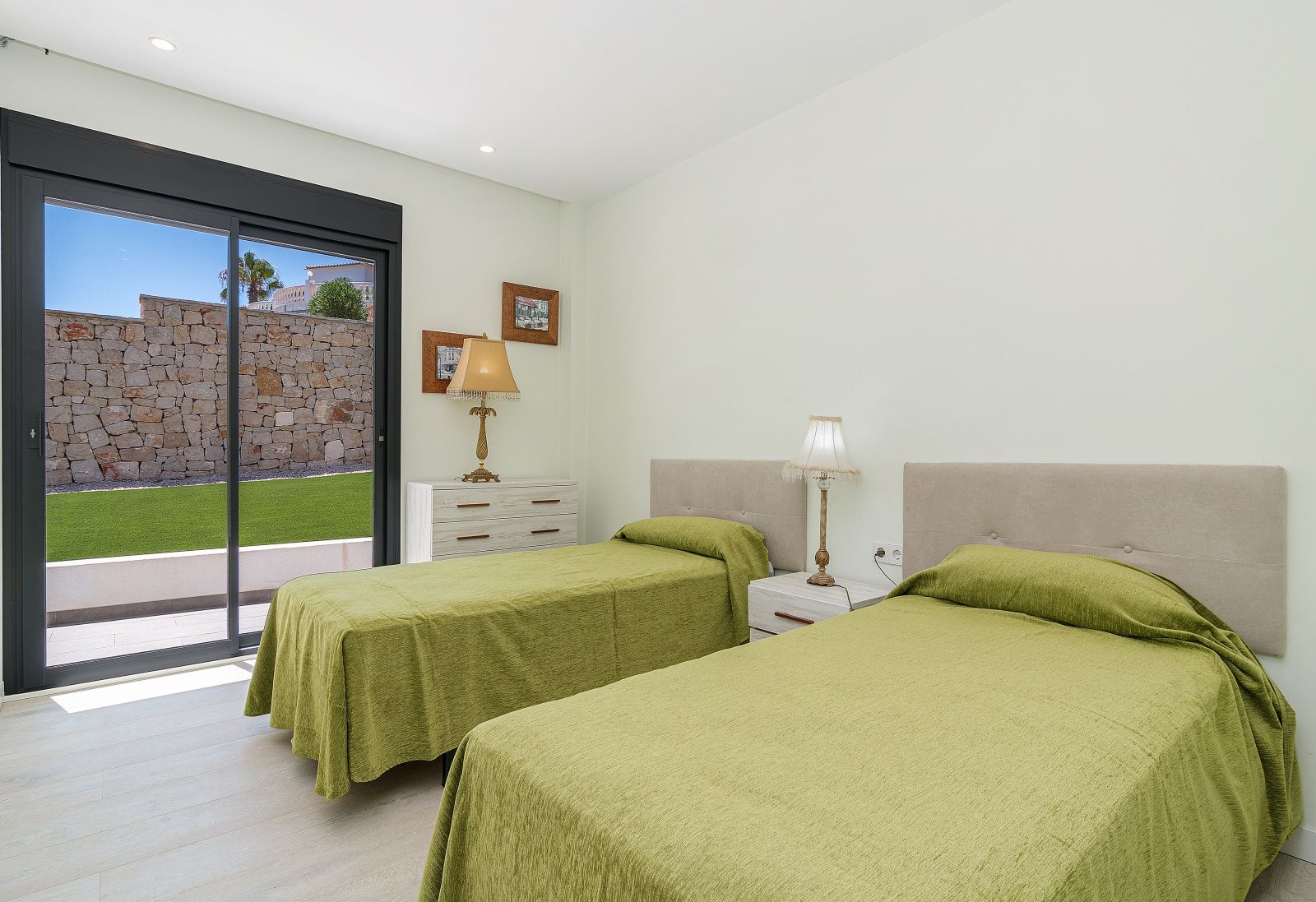 Schlafzimmer mit zwei Einzelbetten, gepolsterten Kopfteilen und direktem Zugang zum Garten durch große Glasschiebetüren. Heller Holzboden und Einbaustrahler.