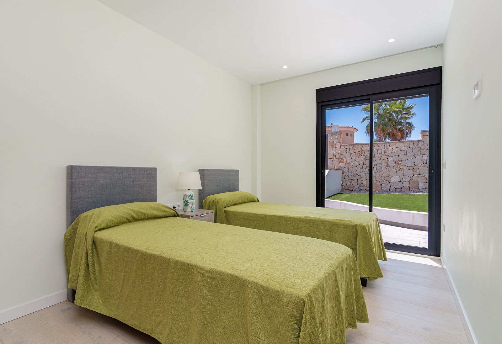 Schlafzimmer mit zwei Einzelbetten, grauen Kopfteilen und direktem Gartenzugang durch große Glasschiebetüren mit schwarzem Rahmen.