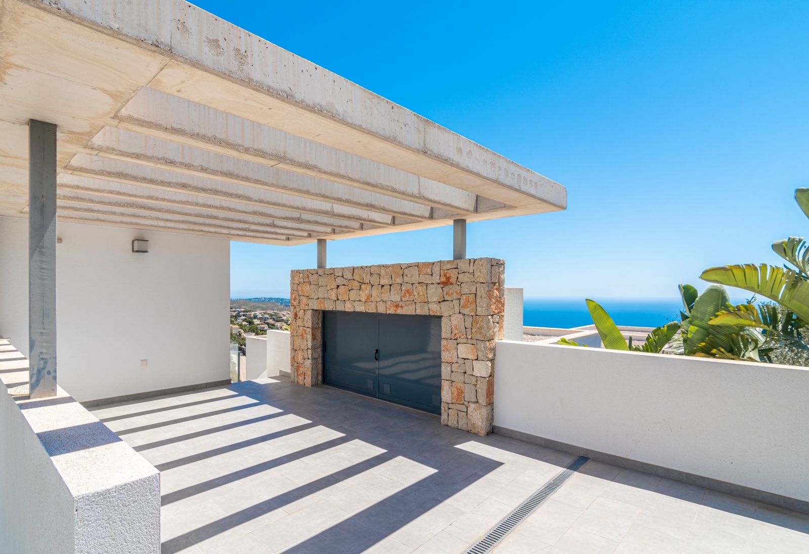 Überdachter Parkplatz mit Sichtbeton-Pergola, Natursteinwand und weitem Blick auf das Mittelmeer und die Küstenlandschaft.