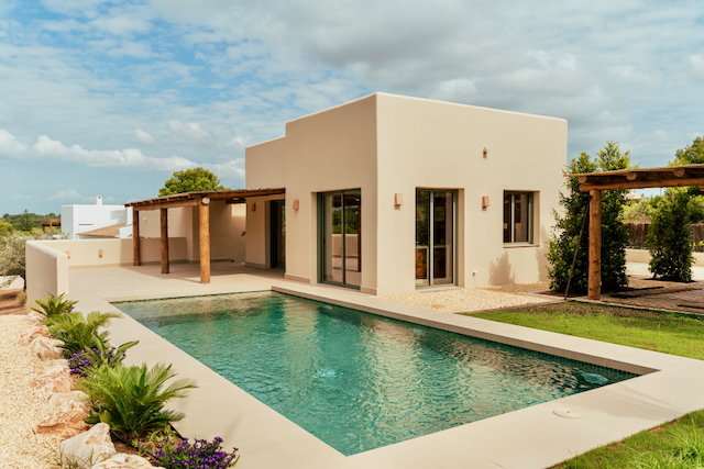 Außenansicht der modernen Villa mit rechteckigem Pool, sandfarbener Fassade und einer rustikalen Holzpergola auf der großzügigen Terrasse.