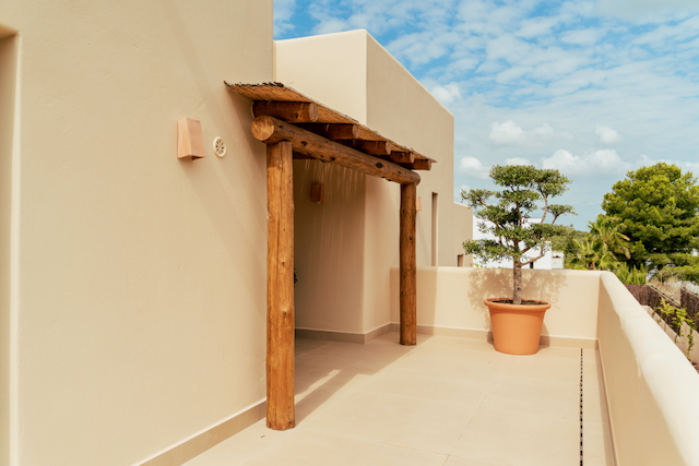 Außenterrasse mit rustikaler Holzpergola, Schilfrohrdach und neutralen Bodenfliesen, ergänzt durch einen dekorativen Baum im Terrakottatopf.