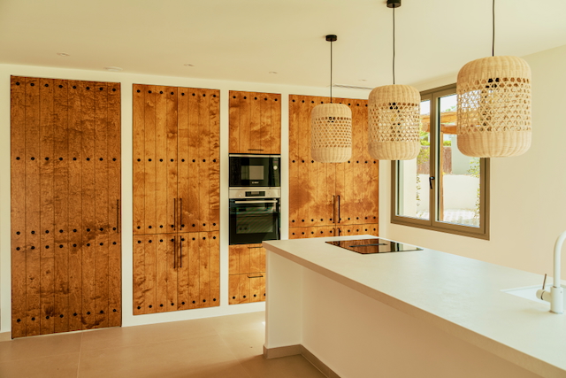 Moderne Küche mit weißer Kochinsel, integrierten Geräten und rustikalen Holzschränken mit Ziernieten. Drei Korblampen sorgen für ein warmes, mediterranes Ambiente.