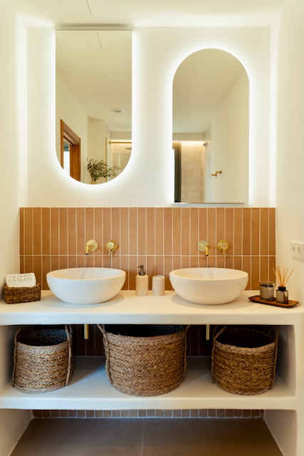 Modernes Badezimmer mit Doppelwaschbecken, goldfarbenen Unterputzarmaturen und hinterleuchteten Bogenspiegeln vor vertikalen terracottafarbenen Fliesen.