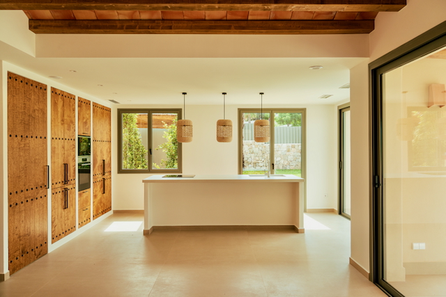 Offene Designerküche mit weißer Kochinsel, rustikalen Holzschränken und Sichtbalkendecke. Große Fensterfronten und Korbpendelleuchten sorgen für ein helles, mediterranes Ambiente.