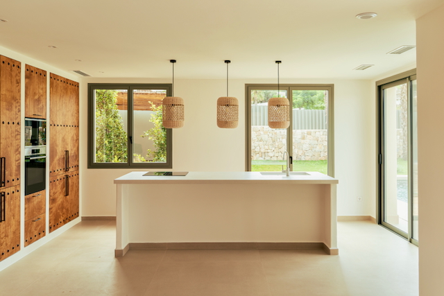 Offene Küche mit weißer Insel, gewebten Pendelleuchten und Holzschränken mit integrierten Geräten. Große Fensterfronten bieten Ausblick auf den Garten und den Poolbereich.